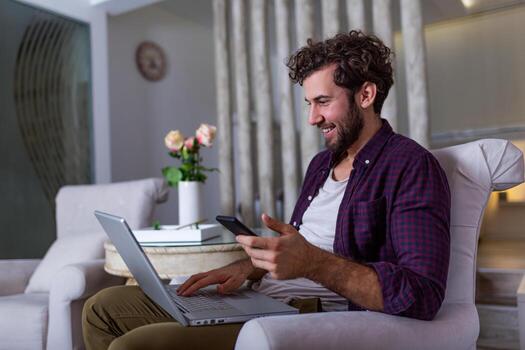 Successful entrepreneur smiling in satisfaction as he checks information on his laptop computer while working in a home office. Young man relaxing on the sofa with a laptop and mobile phone in hands photo