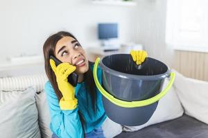 mujer joven preocupada llamando al fontanero mientras el agua de fuga cae en un balde en casa foto