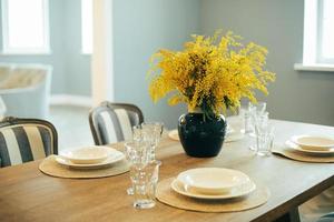 mesa de madera con platos y vasos vacíos. ajuste festivo de la mesa de pascua. ramas de mimosa en florero foto