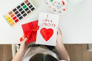 niña sosteniendo en las manos una caja de regalo roja y una tarjeta de felicitación hecha a mano para mamá. concepto del día de la madre foto