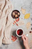 Cup of hot tea in woman's hands. Concrete autumn background with leaves and cozy beige sweater photo