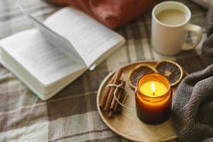 Burning candle with wooden wich in amber glass gar, open book and cup of coffee or tea. Autumn home decor. Cozy fall mood. photo