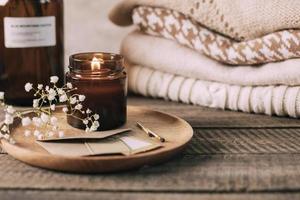 Burning candle in small amber glass jar, flowers of gypsophila and stack knitted seasin sweaters. Cozy lifestyle, hygge concept photo