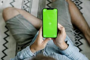 hombre que sostiene un teléfono inteligente móvil con una pantalla verde de clave de croma sentado en el piso en casa foto