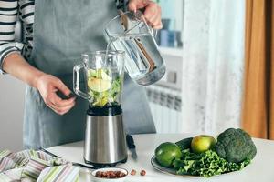 Woman pours water into blender, makes healthy green smoothie. Vegetarian, clean eating diet concept photo