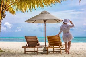 Summer young woman standing backwards and watching beach in white dress, hat. Summer beach colors with girl back view. Summer vacation beach tropical landscape. Bright summer scene, tranquility photo