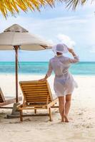 Summer young woman standing backwards and watching beach in white dress, hat. Summer beach colors with girl back view. Summer vacation beach tropical landscape. Bright summer scene, tranquility photo