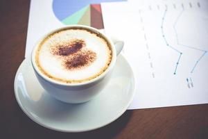 A white coffee cup with a creamy foam placed on the desk with graph data documents. Office work concepts photo