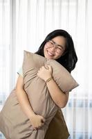 Glasses beautiful Asian woman is hugging her pillow and smiling to the camera in the bedroom with a pastel green - brown color theme. photo