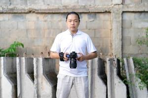 Asian man serious looks at the camera while holds th mirrorless camera medium format in the hand on the construction concret wall background. photo