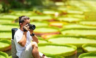 Asian Man is holding a camera and taking a photo at outdoor field with the Lily Lotus Leaf pond background.