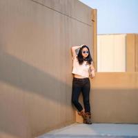 A cute Asian woman stands up and raise elbow up against the wall and wears blue sunglasses at the rooftop of the building at sunset time. photo