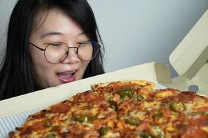 una joven asiática con anteojos abre una caja de pizza y se emociona con la pizza de gran tamaño que contiene. disparar con luz de estudio. foto