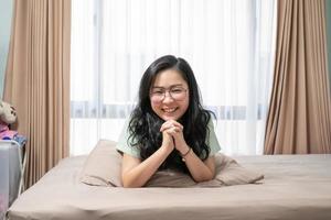 Beautiful Asian woman hold her hands together while lie on her stomach on bed in bedroom with a pastel green - brown color theme. photo
