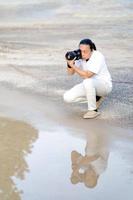 el hombre asiático sostiene y mira seriamente dentro del tipo de formato medio de la cámara sin espejo del visor en el suelo de asfalto de cemento frente al reflejo del agua. foto