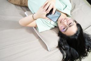 hermosas gafas mujer asiática yacen boca arriba, charlando, escribiendo y enviando mensajes de texto, móvil en su dormitorio con un tema de color verde pastel - marrón.