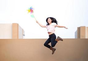 una mujer asiática publica, juega y salta con un juguete de molino de viento de color arcoíris en la azotea del edificio al atardecer. foto