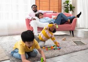 niños felices jugando juguetes y disfrutando de la actividad de juego mientras se sientan en la alfombra del piso en la sala de estar. pareja negra de padres descansando en el sofá y viendo a los niños foto