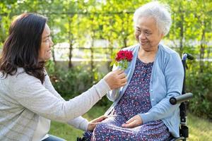 Caregiver daughter hug and help Asian senior or elderly old lady woman holding red rose on wheelchair in park. photo