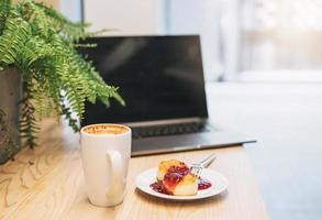 Open laptop, cheesecakes with jam, cup of cappuccino on the table in cafe photo