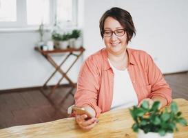 retrato de mujer de negocios morena encantadora adulta con gafas de cuerpo positivo de tamaño grande usando el móvil en la oficina del hogar foto