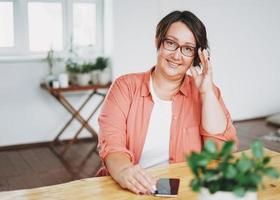 retrato de mujer de negocios morena encantadora adulta con gafas de cuerpo positivo de tamaño grande usando el móvil en la oficina del hogar foto