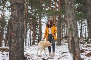 Young smiling woman in yellow jacket with big kind white dog Labrador walking in winter forest photo