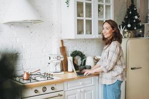 atractiva mujer sonriente con el pelo rizado en camisa a cuadros con portátil cerca de la ventana en la cocina luminosa en casa foto