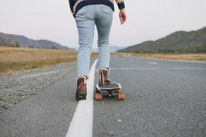 Crop photo of women's legs in jeans on longboard on road with the mountains background