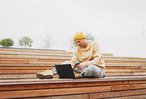 Stylish man hipster in yellow hat freelancer working on laptop in the street city park photo