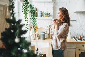 atractiva mujer sonriente con el pelo rizado en camisa a cuadros con una taza de té cerca de la ventana en la cocina luminosa en casa. tiempo de Navidad foto