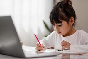 Asian schoolgirl doing her homework with laptop at home. Children use gadgets to study. Education and distance learning for kids. Homeschooling during quarantine. Stay at home photo