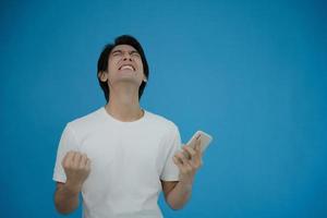 Cool asian man excited and surprised by smartphone on blue background. A man saw a sale on an online shopping website or See the results of the football team cheering competition. photo