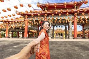 Rear view of Asian woman in red cheongsam qipao dress is holding her lover hand while visiting the Chinese Buddhist temple during lunar new year for traditional culture concept photo