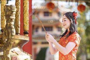 Asian woman in red cheongsam qipao dress is offering incense to the ancestral god inside Chinese Buddhist temple during lunar new year for  for best wish blessing and good luck concept photo