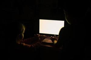 Kids tap on laptop while watch on colored screen in light off room. photo