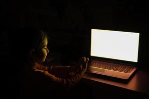 la niña toca en la computadora portátil mientras mira en la pantalla a color en la habitación sin luz. foto