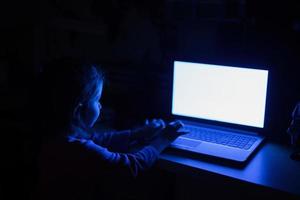 la niña toca en la computadora portátil mientras mira en la pantalla a color en la habitación sin luz. foto