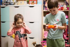 Brother hold remote control and sister listening to music through wired headphones in children's room. photo