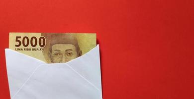 Indonesian rupiah banknotes worth IDR 5,000 in a white envelope isolated on red background. Conceptual business illustration photo