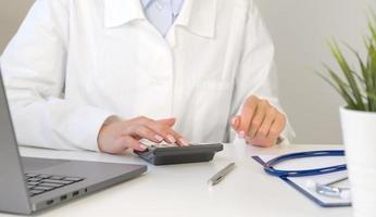 Front view at unrecognizable doctor using calculator with laptop and stethoscope near. Medical insurance concept photo