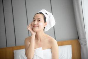 Happy young Asian woman applying face lotions while wearing a towel and touching her face. Daily makeup and skincare photo