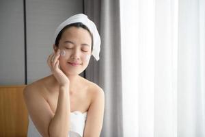 Happy young Asian woman applying face lotions while wearing a towel and touching her face. Daily makeup and skincare photo