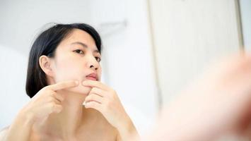 piel problemática. Mujeres asiáticas jóvenes preocupadas haciendo estallar granos en la mejilla mientras están de pie cerca del espejo en el baño. jóvenes asiáticas con acné