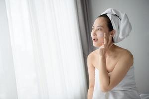 feliz joven asiática aplicando lociones faciales mientras usa una toalla y toca su cara. maquillaje diario y cuidado de la piel foto