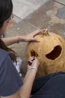 female carving large orange pumpkin for Halloween while sitting at wooden table at home photo