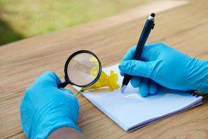Student  hands in protective gloves hold magnifying glass to inspect flowers, holds pen to write on paper. Concept, science subject, project work. Experiment, education, learning by doing approach. photo