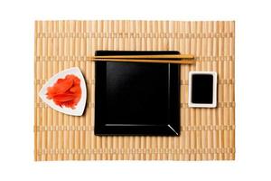 Empty black square plate with chopsticks for sushi, ginger and soy sauce on yellow bamboo mat background. Top view with copy space for you design photo