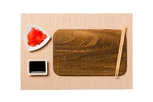 Empty rectangular brown wooden plate with chopsticks for sushi, ginger and soy sauce on brown sushi mat background. Top view with copy space for you design photo
