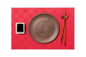 Empty round brown plate with chopsticks for sushi and soy sauce on red mat sushi background. Top view with copy space for you design photo